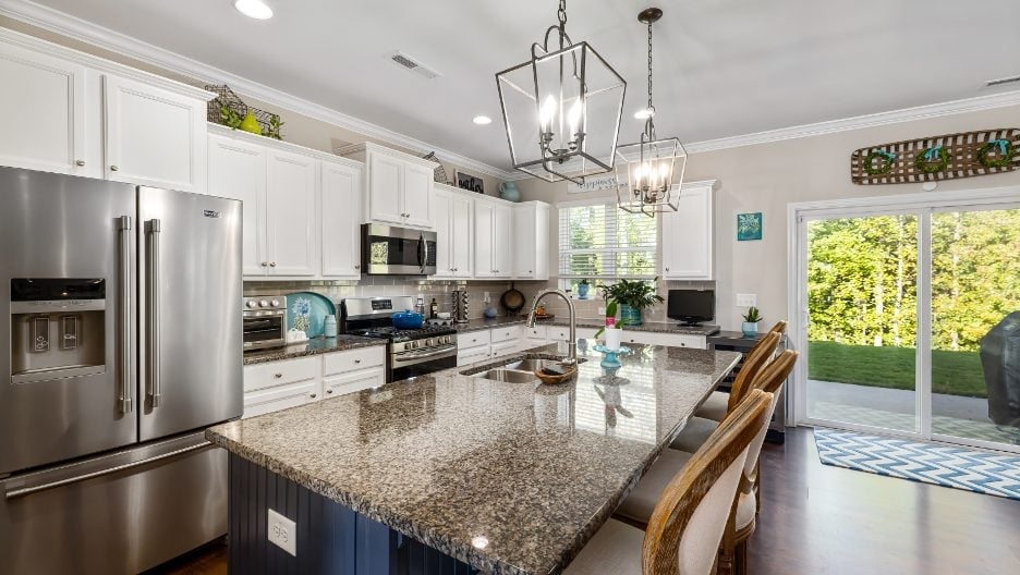 A kitchen with a large island and stainless steel fridge. The kitchen has white cabinets and a view of the outdoors.