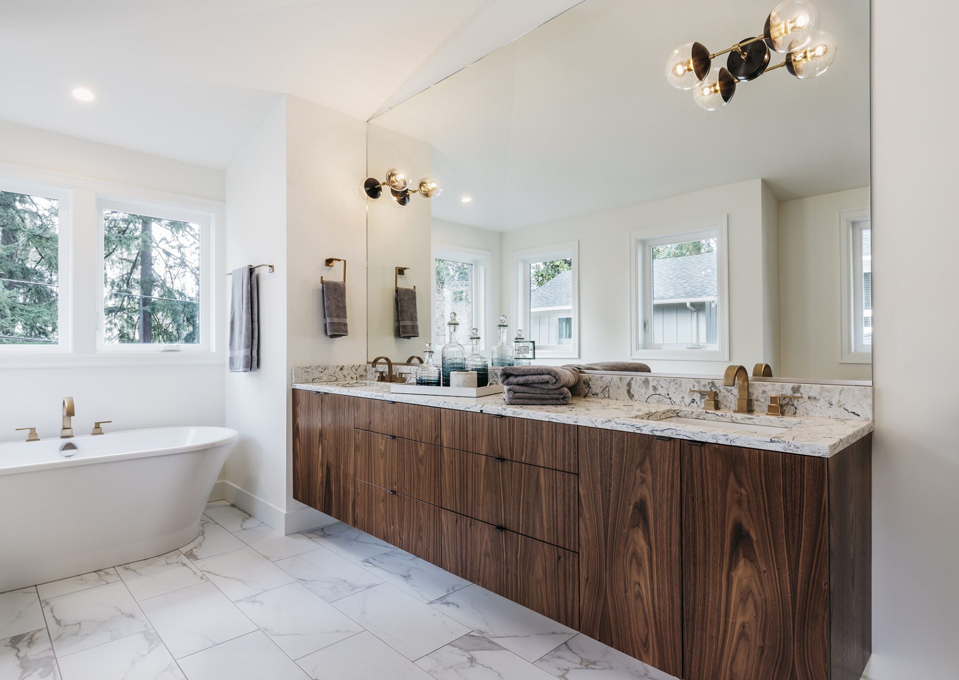 A modern bathroom with a large double vanity and a spacious mirror. The room features a bathtub and windows with natural light.