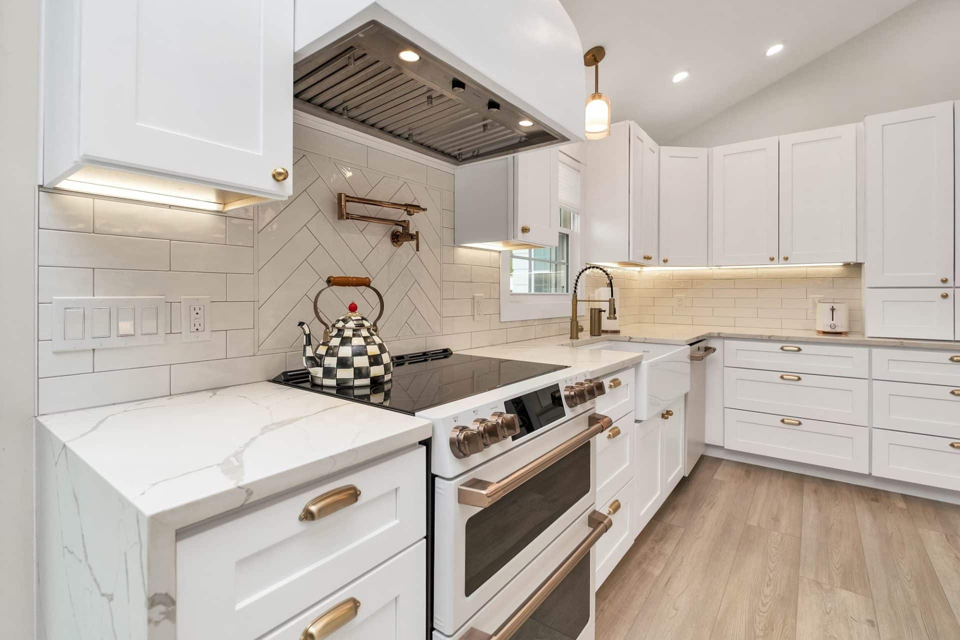 A modern kitchen with white cabinets and gold hardware. The kitchen features a white marble countertop and a black cooktop.