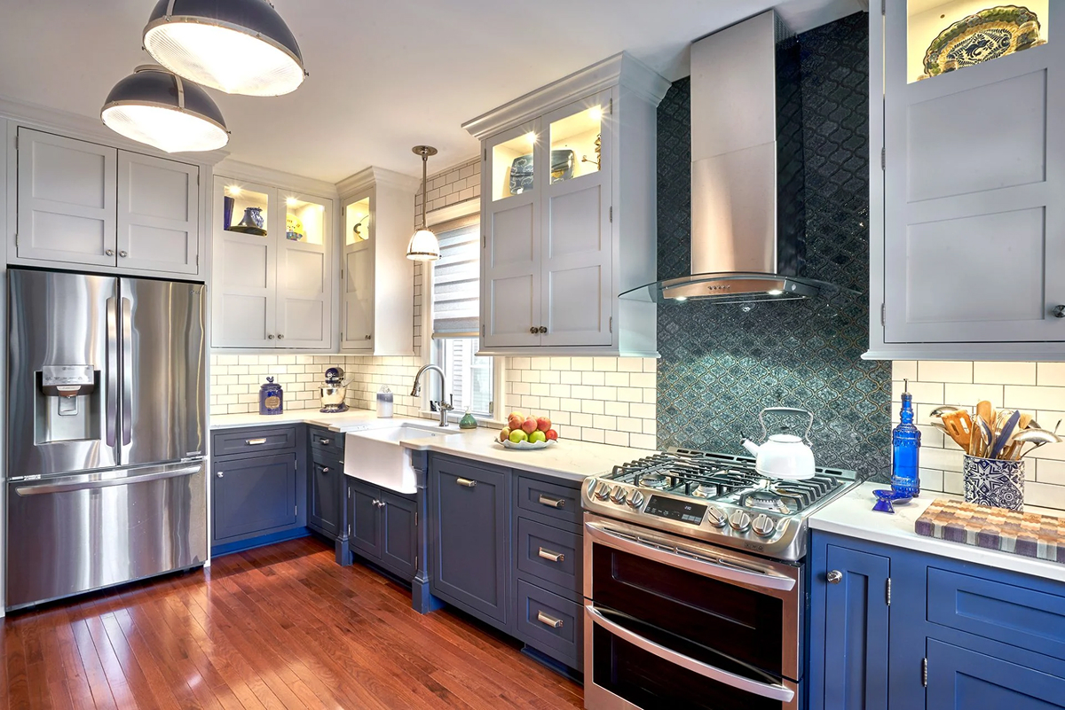 A modern kitchen with blue lower cabinets and stainless steel appliances. The kitchen features a white countertop and a green backsplash.