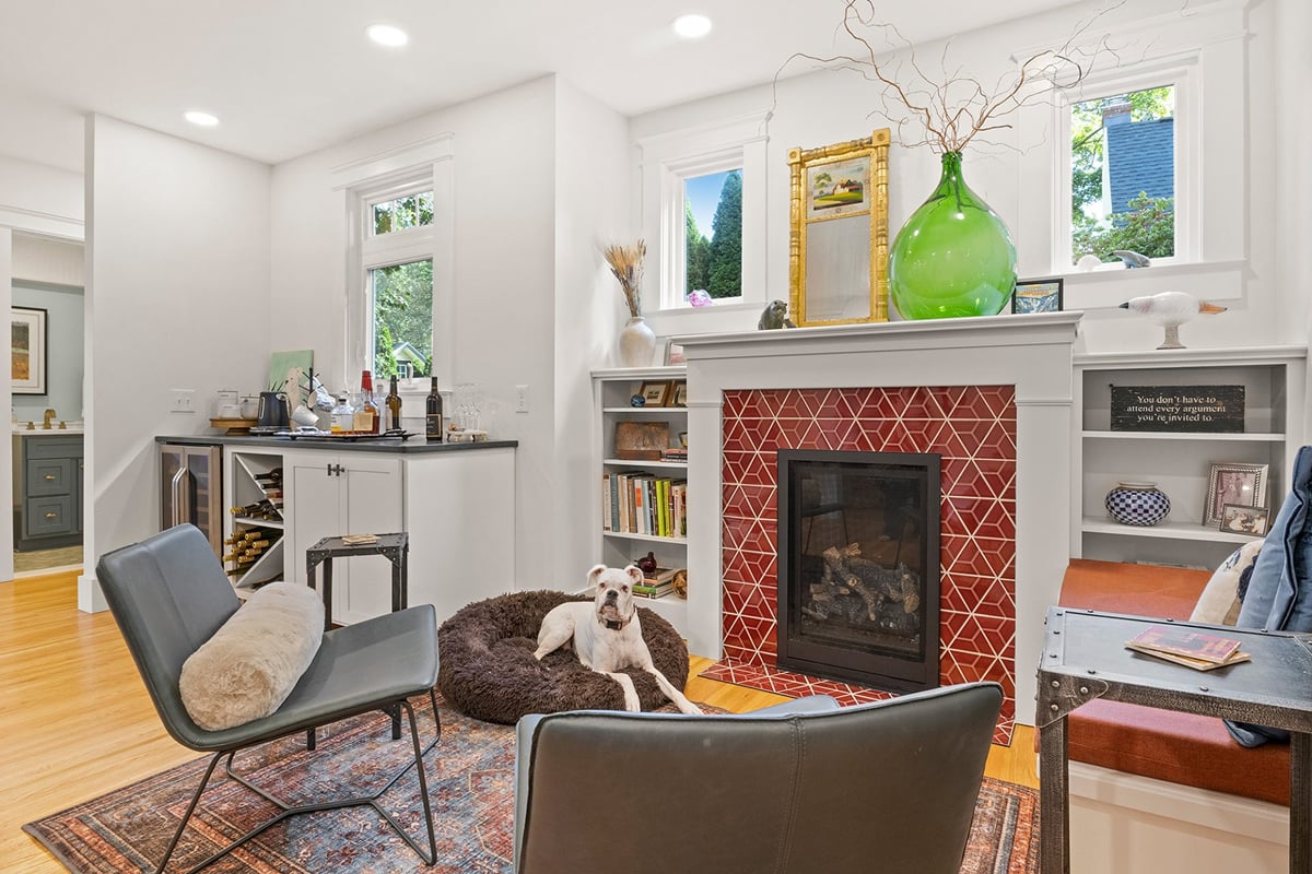 A dog lying on a dog bed in front of a fireplace. The room is furnished with a chair and a table.