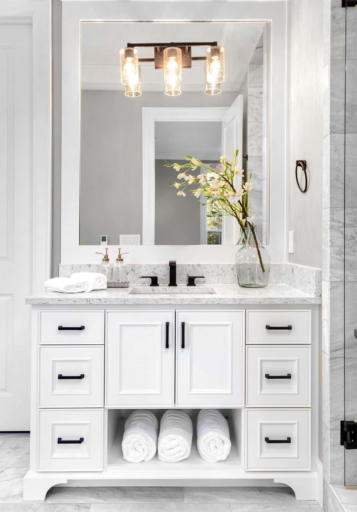 A clean and elegant bathroom vanity with a large mirror and a vase of flowers. The vanity features multiple drawers and a marble countertop.