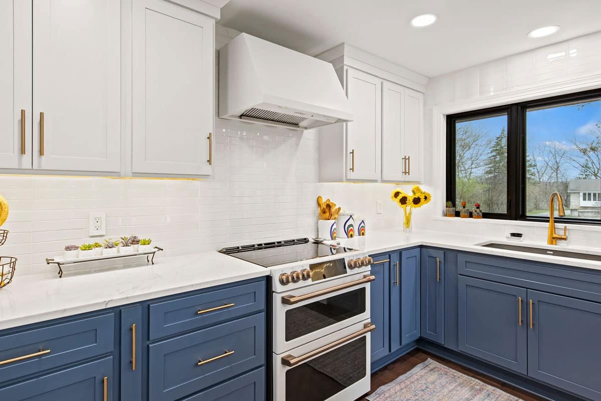 Modern kitchen interior with blue lower cabinets, white upper cabinets, and white countertops. The kitchen features a stainless steel oven and range hood.