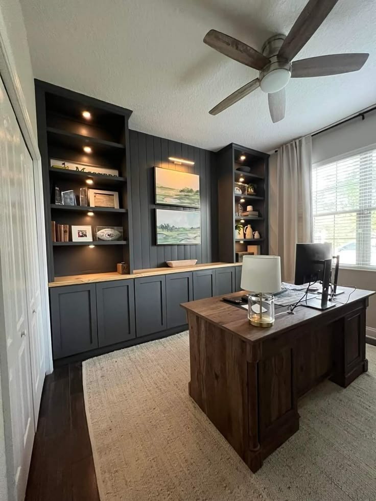 A modern home office with a large wooden desk, a ceiling fan, and built-in bookshelves. The room has a window with a view.