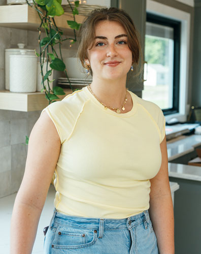 A young woman wearing a yellow shirt and jeans standing in a kitchen.