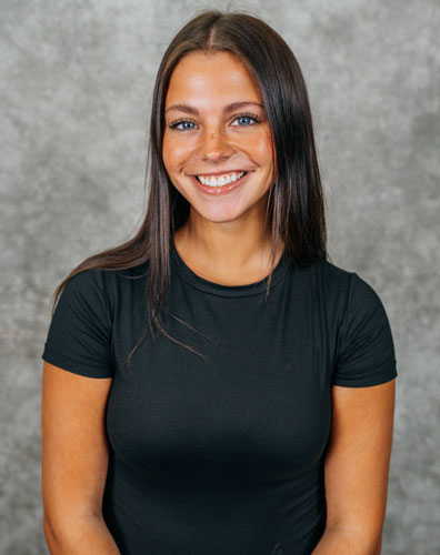 A young woman with long dark hair and a black t-shirt smiles at the camera. She has a warm and friendly expression.