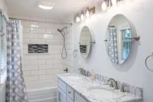 A stylish bathroom featuring a white subway tile shower, a marble countertop vanity with dual sinks, two round mirrors, and a patterned gray shower curtain.