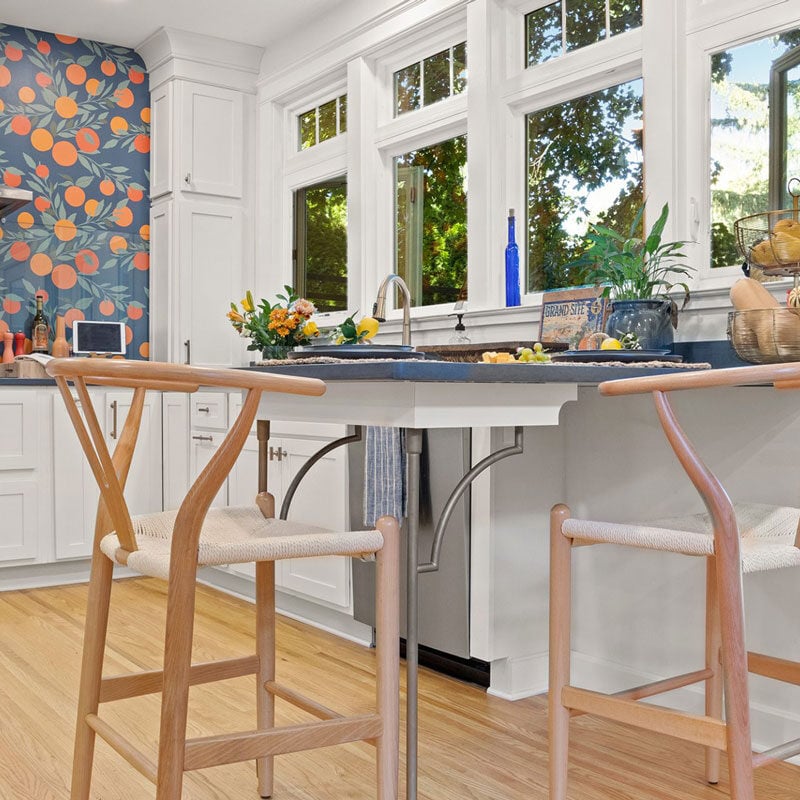 Modern kitchen island with two stools and fruit bowl. Kitchen has colorful backsplash and large windows.
